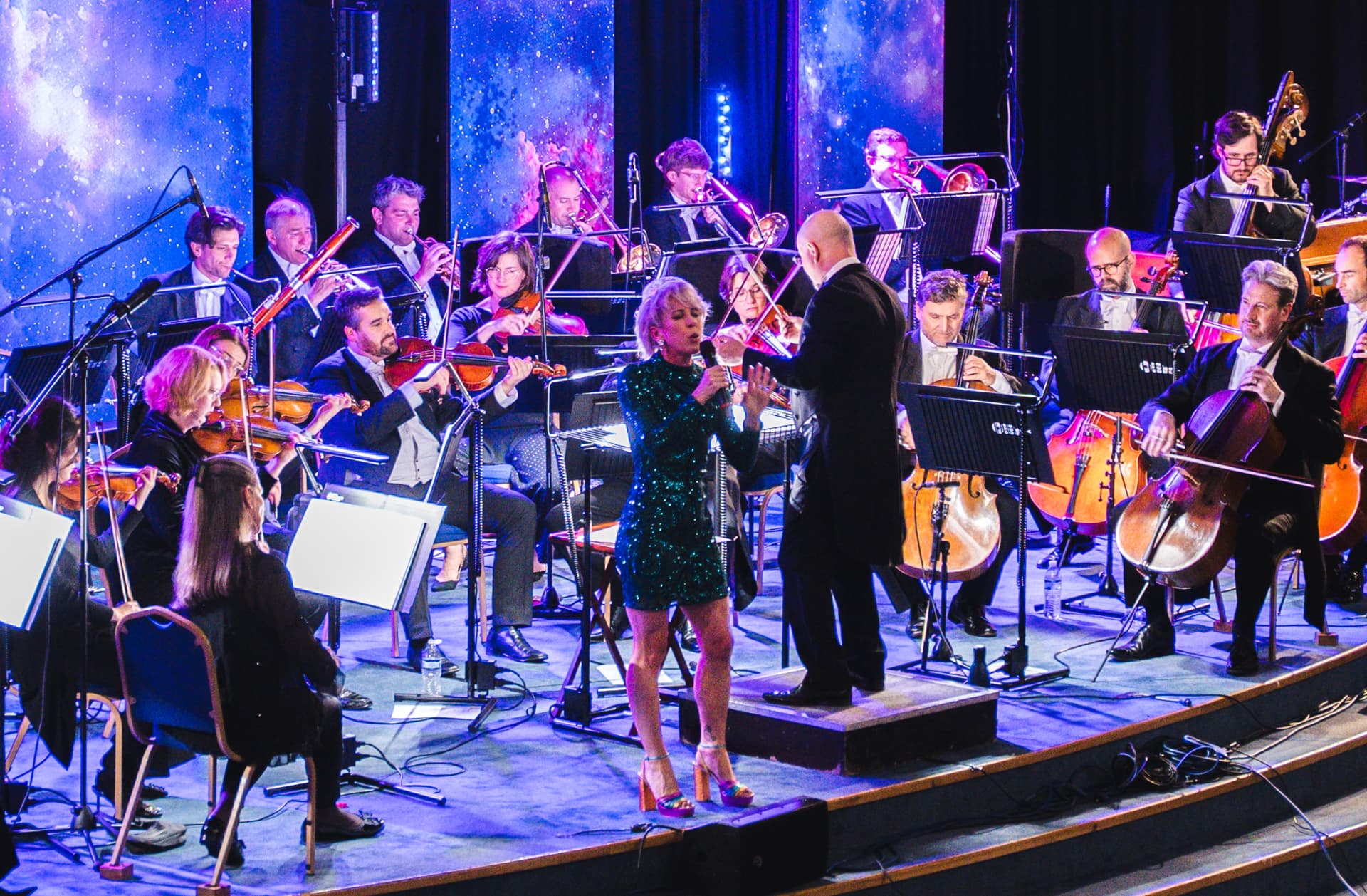 Orchestra performing on stage with a singer in a green dress beside the conductor